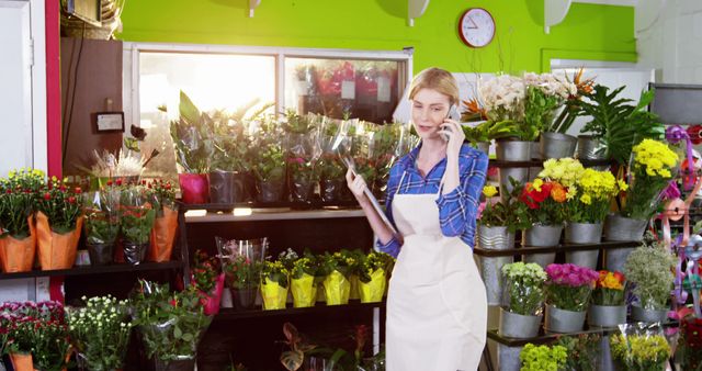 Florist wearing apron arranging flowers and on a phone call in flower shop full of colorful flowers and plants. Perfect for business concept images, retail industry promotions, small business advertising, or floral arrangement guides.