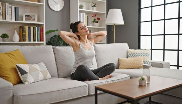 Calm woman stretching on modern sofa in bright living room practicing gentle home wellness