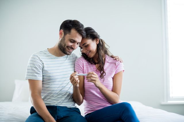 Couple sitting on bed, smiling and embracing while looking at pregnancy test. Ideal for use in articles or advertisements related to pregnancy, family planning, parenthood, and home life. Can also be used in blogs or social media posts celebrating pregnancy announcements and family moments.
