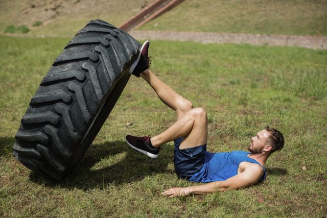 This image shows a fit man performing a leg workout with a large tire in an outdoor boot camp setting. Ideal for use in fitness blogs, workout guides, advertisements for fitness programs, and promotional materials for outdoor training camps.