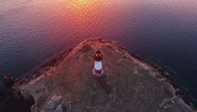 Red and White Lighthouse Glowing at Sunset on Rocky Headland Aerial Seascape