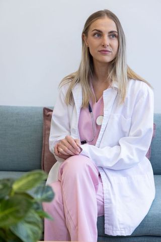 Female clinician sitting in minimalist clinic lounge wearing pink scrubs and white coat