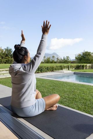 Asian woman practicing yoga on patio by pool in serene backyard, stretching and meditating