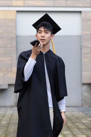 Asian graduate speaking into smartphone wearing cap and gown on campus courtyard