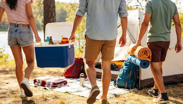 Friends heading to lakeside campsite carrying cooler and camping gear on sunny day
