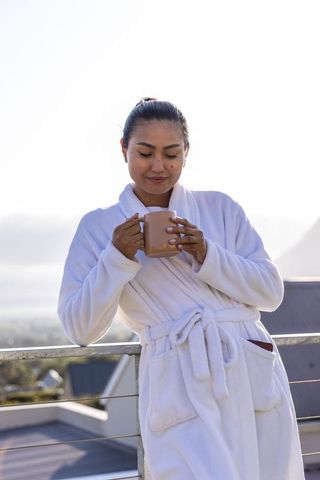 Asian woman in white plush robe sipping morning coffee on rooftop balcony at sunrise