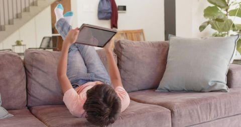 Indian preteen lying upside-down on sofa using tablet for relaxed entertainment at home