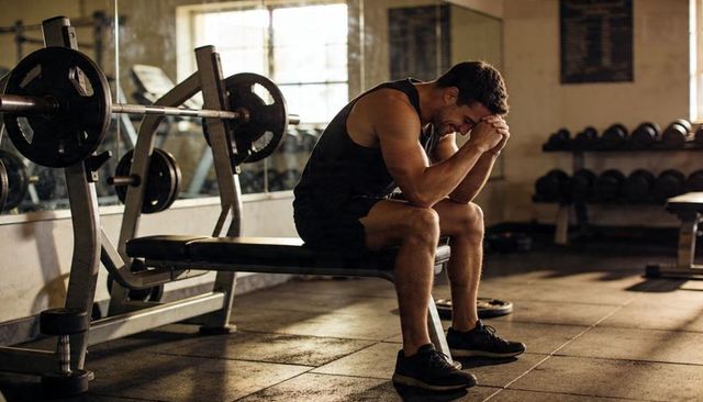 Muscular man resting on bench after intense weightlifting in industrial gym with loaded barbell