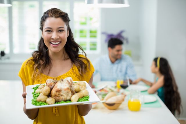 Smiling woman holding tray of roasted chicken and vegetables in a bright kitchen. Family in the background enjoying a meal together. Perfect for use in advertisements, blogs, or articles about family meals, cooking, healthy eating, and domestic life.