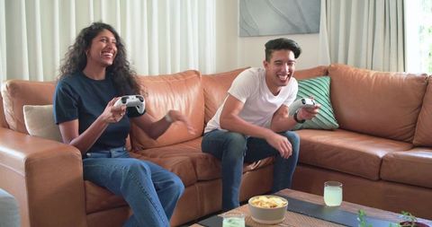 African american couple playing video games on leather sofa laughing together at home