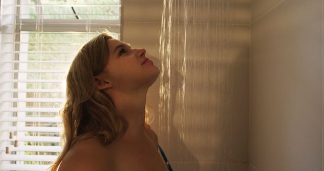 Young woman with blonde hair enjoying a shower, with sunlight streaming in through a nearby window. Can be used for topics related to hygiene, self-care, relaxation, bathing routines, and personal well-being. Suitable for lifestyle, health, and beauty contexts.