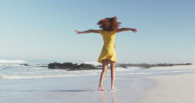 Woman standing on sandy beach with arms outstretched, wearing yellow dress, enjoying sunny day. Perfect for travel advertisements, vacation brochures, lifestyle blogs, outdoor activity promotions.