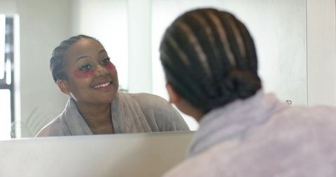 African american woman applying red under-eye patches at bathroom mirror for skincare ritual