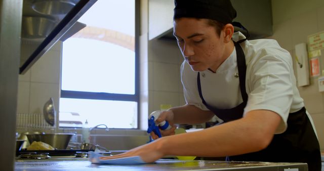 Young chef wearing uniform cleaning kitchen counter with spray bottle in restaurant kitchen. Perfect for websites, blogs, or articles about restaurant hygiene, culinary professions, food safety practices, and clean working environments.