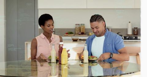 Couple preparing fresh salads at glass table with condiments in modern kitchen