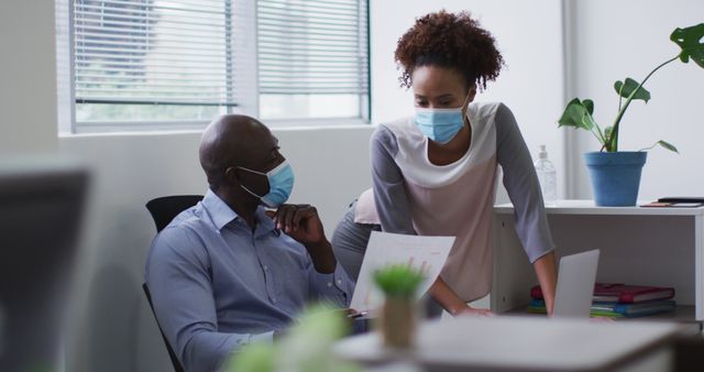 Two colleagues wearing face masks are engaged in discussion in a modern office setting. A man is seated, while a woman is standing, holding a piece of paper with charts. There are elements of office supplies, plants, and modern furniture indicating a professional workspace. Useful for conveying pandemic-related workplace scenarios, business communication, teamwork, and health safety measures in a corporate environment.