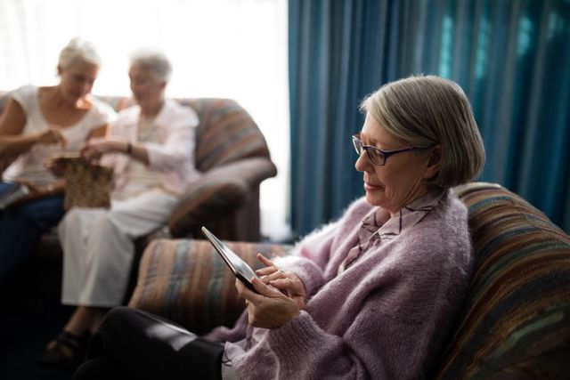 Senior woman sitting on an armchair using a digital tablet in a retirement home. Two other elderly women are sitting on a couch in the background, engaging in a different activity. This image can be used for promoting elderly care facilities, technology use among seniors, and independent living for the elderly.