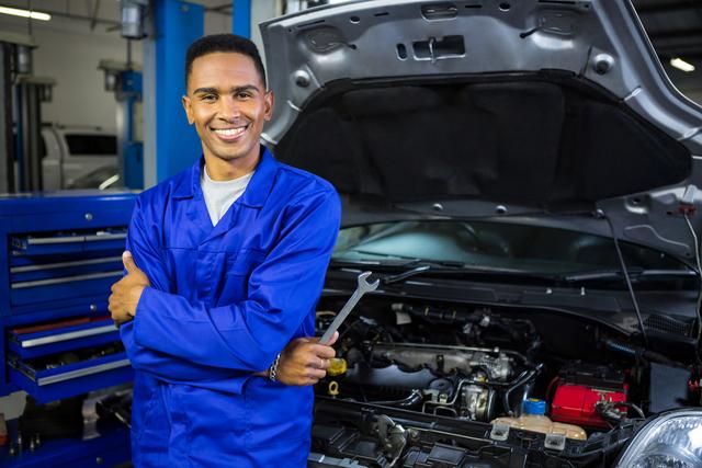 Male mechanic in blue overalls holding spanner, standing confidently in front of car with open hood in repair garage. Perfect for automotive repair services, engineering professions, and mechanical workshops advertisements. Can be used to promote skilled labor, customer service excellence, and professional mechanic services.