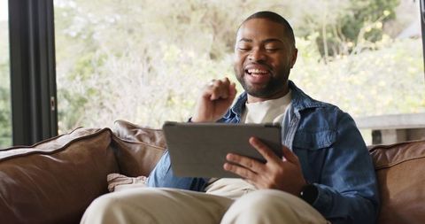 African American man smiling on couch using tablet, celebrating good news at home