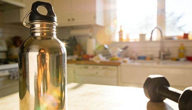 Sunlit stainless water bottle reflecting person holding phone, dumbbell on kitchen countertop
