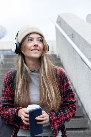 Young woman commuter with headphones holding coffee and smartphone on urban stairs