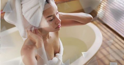 Mid-adult woman adjusting towel turban in sunlit oval bathtub for serene self-care moment