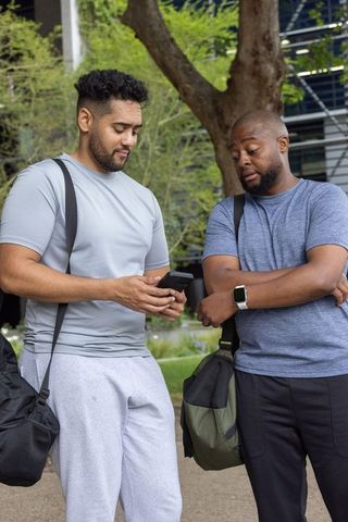 Two african american men in sportswear sharing smartphone, carrying gym duffel bags in urban plaza