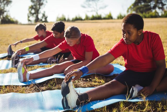 Group of kids stretching on blue mats during an outdoor boot camp on a sunny day. They are wearing red shirts and black shorts, focusing on their flexibility and fitness. Ideal for use in articles about children's health, fitness programs, outdoor activities, teamwork, and summer camps.
