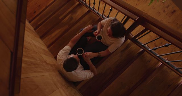 This image captures an overhead view of two men sitting on stairs, sharing cups of coffee. The scene takes place on a wooden floor, creating a warm and cozy atmosphere. It can be used to depict themes of friendship, relaxation, casual conversation, and indoor bonding moments. Ideal for websites related to coffee shops, friendship, lifestyle blogs, and advertisements promoting coffee or living spaces.