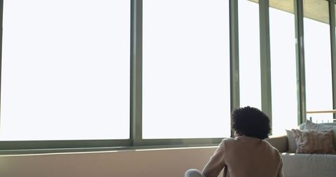 African american man sitting on sofa by floor-to-ceiling windows in modern minimalist loft