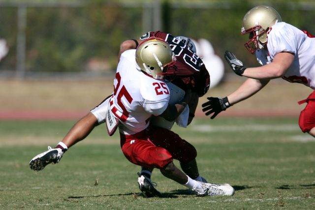 This image captures a moment of intense tackle during a football game on a grass field. Football players in red and white uniforms are engaged in physical contact, showcasing the dynamic and competitive nature of the sport. Ideal for use in sports articles, athletic training materials, promotional content for football events, and illustrating teamwork concepts.