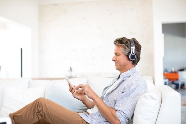 Man sitting on couch in living room, wearing headphones and using mobile phone. Ideal for concepts related to home entertainment, relaxation, modern technology, and casual lifestyle.