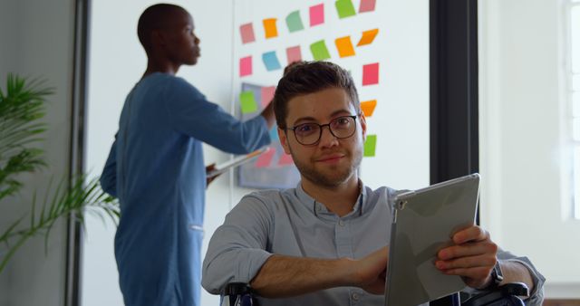 Young professional in wheelchair using tablet with colleague in background brainstorming - Download Free Stock Images Pikwizard.com