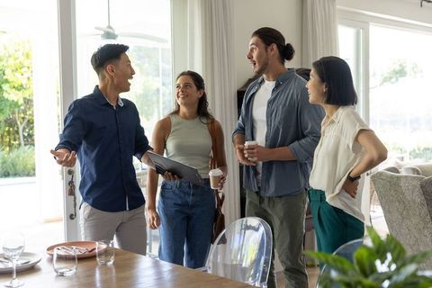 Diverse friends chatting and touring modern dining room holding tablet and coffee cups