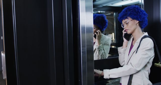 A young professional woman with a stylish blue afro hairstyle is using her phone while standing in an elevator. She is dressed in business attire, including a white jacket, and wearing glasses, giving a serious look. This image can be used to depict themes related to urban lifestyle, corporate environment, business communications, and contemporary fashion.