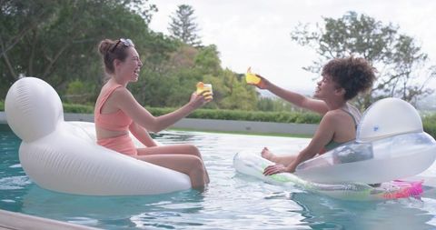Friends clinking drinks on pool floats enjoying sunny backyard summer relaxation