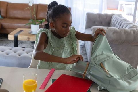 African girl packing mint-green backpack at table with school supplies