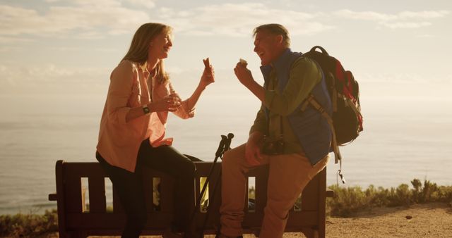 This image features a senior couple sitting on a bench, enjoying snacks during a hike by the ocean. The setting sun casts a warm glow, enhancing the scenic view of the sea in the background. Ideal for articles or marketing materials on outdoor activities, healthy living, senior adventures, and retirement lifestyle.
