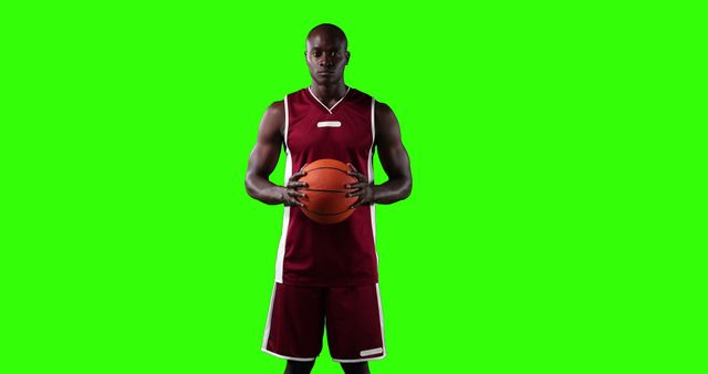 Basketball player wearing red uniform holding basketball against green screen background, showing strength and focus. Ideal for sports promotions, fitness campaigns, athletic advertisements, and visual effects requiring diverse background replacements.