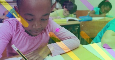 Black schoolgirl writing at desk concentrating on homework in classroom with colorful overlays