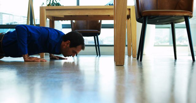 Businessman in blue shirt doing push-ups under desk in modern office. Great for promoting workplace fitness programs, healthy lifestyle in corporate environments, balancing work and physical activity.