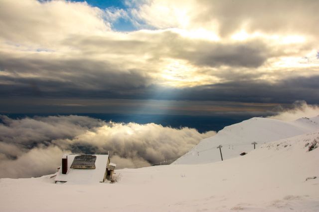 Panoramic view of a snow-covered mountain with a blend of clouds and sunrays breaking through. An isolated ski lodge is nestled in the snow, evoking feelings of solitude and serenity. Ideal for use in travel and tourism publications, advertisements for winter sports, or inspirational content.