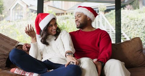 Diverse couple wearing santa hats sitting on sofa showing smartwatch in cozy holiday room