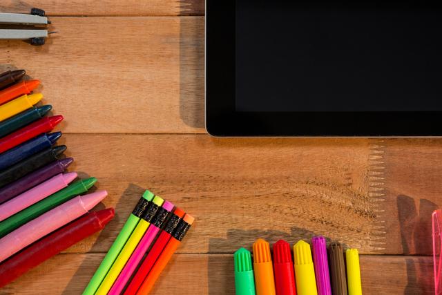 Close-up view of a digital tablet surrounded by colorful pencils, pens, and markers on a wooden table. Ideal for illustrating concepts of modern education, creative workspace, or back-to-school themes. Suitable for use in educational blogs, tech articles, or creative project advertisements.