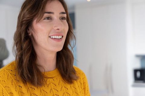 Smiling woman in mustard yellow knit sweater in modern white kitchen, cozy home portrait