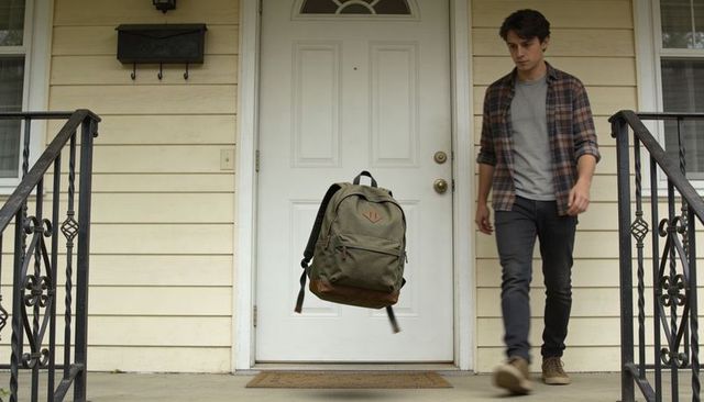 Olive green backpack floating before white door with young man walking on porch