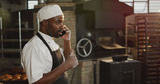 Baker on phone in bakery kitchen surrounded by pastries and industrial baking equipment. Ideal for business communications, bakery industry, food production, small business operations, and culinary professions.