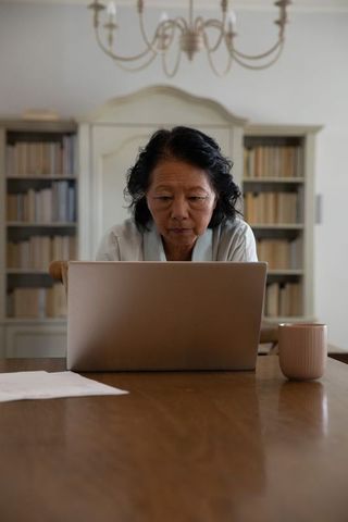 Senior chinese woman working on laptop at wooden dining table with books, notes and mug at home