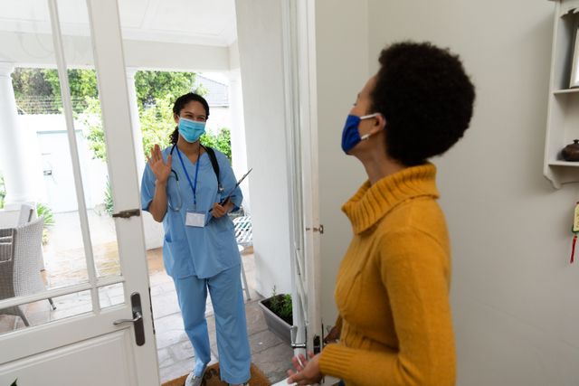 Female doctor in blue scrubs and face mask greeting a woman at her door. Both individuals are wearing face masks, emphasizing safety and hygiene during the COVID-19 pandemic. Ideal for use in articles or advertisements related to healthcare, home visits, pandemic safety measures, and patient care.