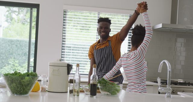 Young African American couple joyfully cooking together in a modern kitchen. They are dancing and smiling, enjoying their time while preparing a meal. The scene is filled with natural light from large windows. Perfect for themes surrounding love, relationships, healthy living, and modern home life.